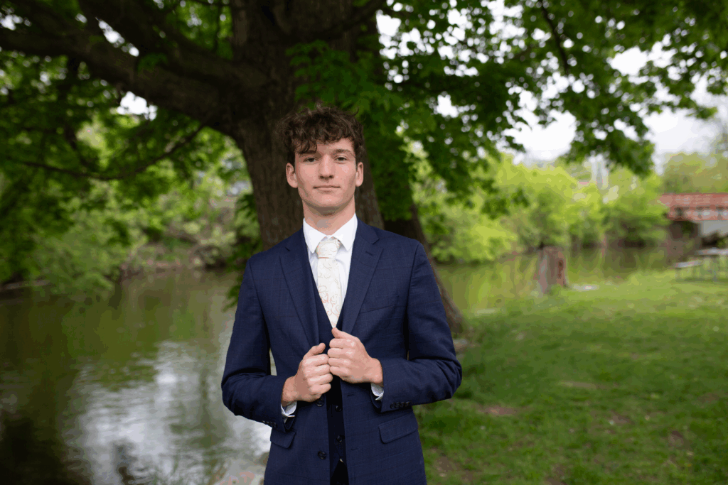 Handsome young man with dark curly hair wearing a dark blue suit, white shirt, and standing in front of a river